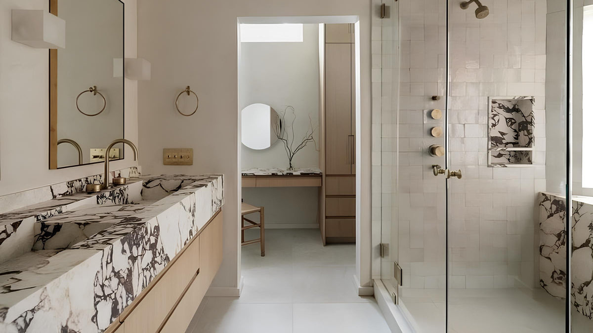 A modern bathroom interior with a double vanity featuring black-and-white marble sinks and countertop, a large wall mirror, a walk-in shower with glass doors, and light wood cabinetry.