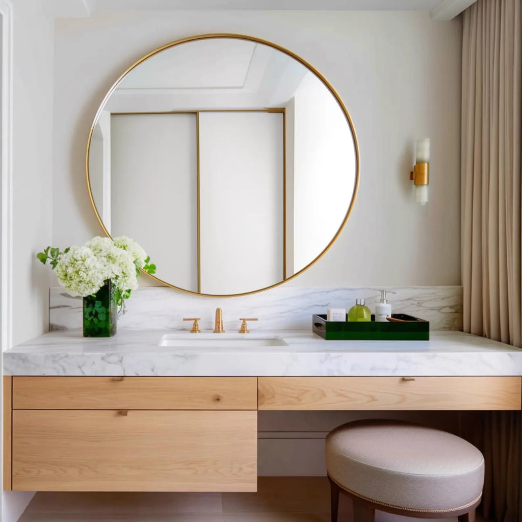 A modern bathroom vanity setup featuring a large round gold-framed mirror, a white marble countertop, light wood drawers, a green tray with toiletries, and a round upholstered stool.