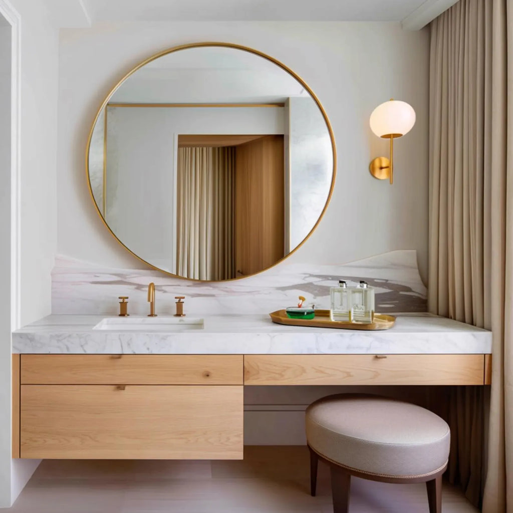 A modern bathroom vanity setup with a large round gold-framed mirror, white marble countertop, light wood floating cabinet, brass faucet, wall sconce, and a round upholstered stool in a softly lit interior.