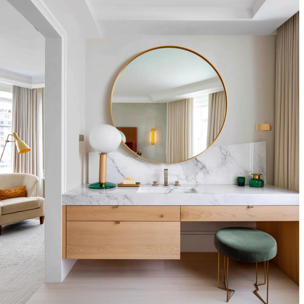 A modern bathroom vanity setup featuring a large round gold-framed mirror, a white marble countertop with an integrated sink, light wood drawers, a green velvet stool, and decorative accessories.