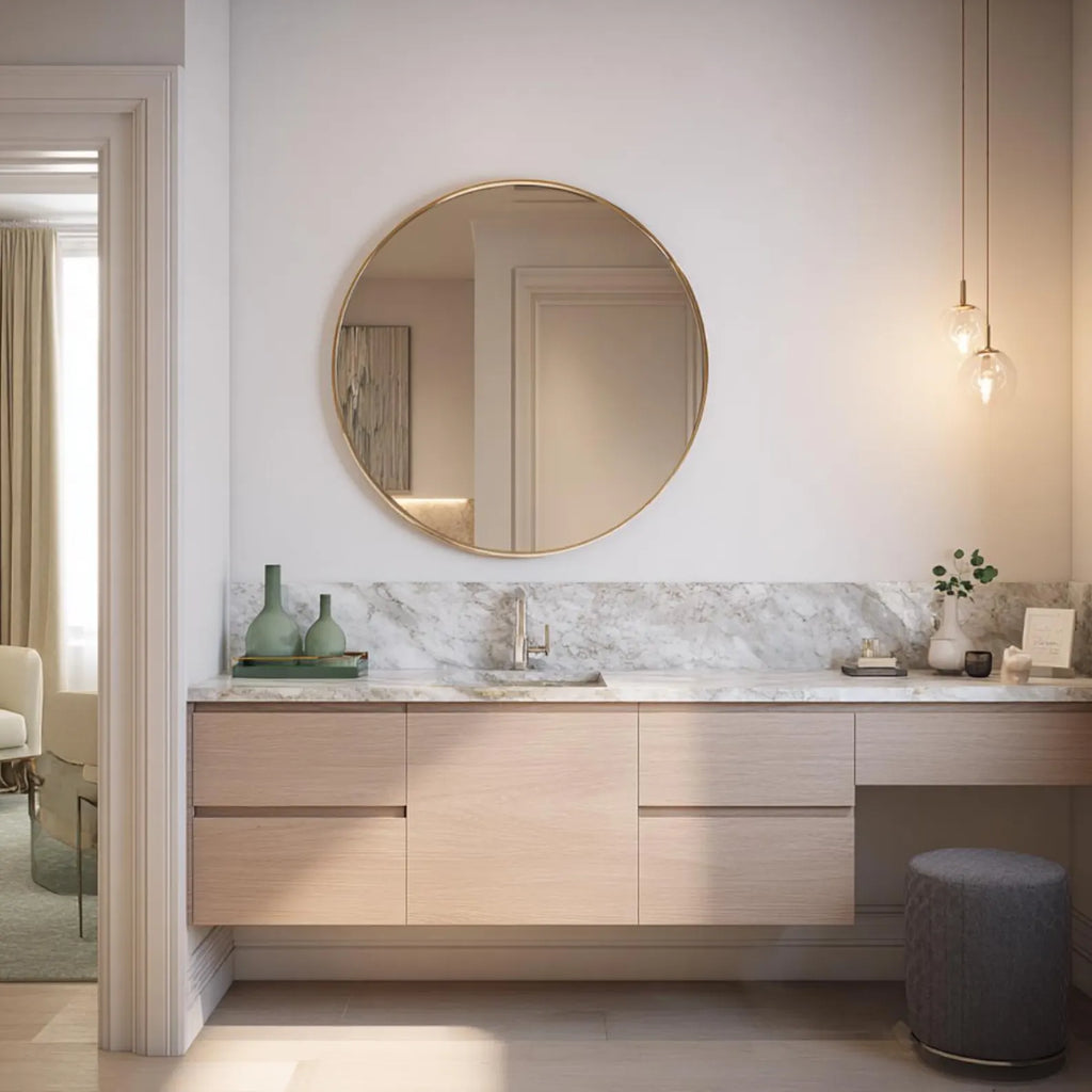 A modern bathroom vanity setup featuring a large round gold-framed mirror mounted on a white wall above a light wood floating cabinet with a marble countertop. The scene includes decorative green vases, a chrome faucet, pendant lights, and a gray upholstered stool.