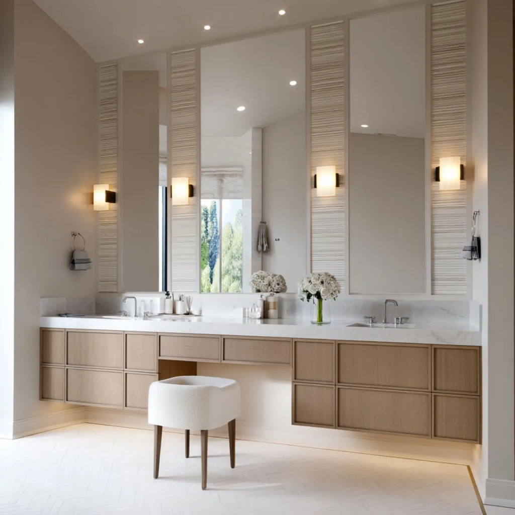 A modern double-sink bathroom vanity with light wood cabinets, a white marble countertop, large wall mirrors, and a white upholstered stool in a bright, neutral-toned bathroom.