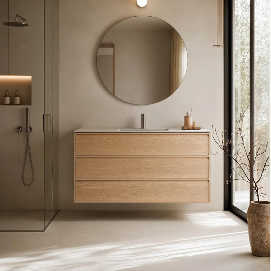 A modern floating bathroom vanity with three light oak wood drawers, a white integrated sink, and a chrome faucet, set against a beige wall beneath a round wall mirror. A glass shower enclosure is visible on the left, and a large window with a vase of branches is on the right.