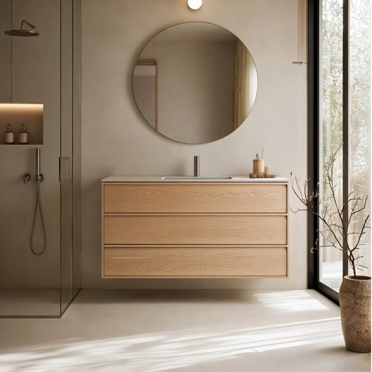 A modern floating bathroom vanity with three light oak wood drawers, a white integrated sink, and a chrome faucet, set against a beige wall beneath a round wall mirror. A glass shower enclosure is visible on the left, and a large window with a vase of branches is on the right.