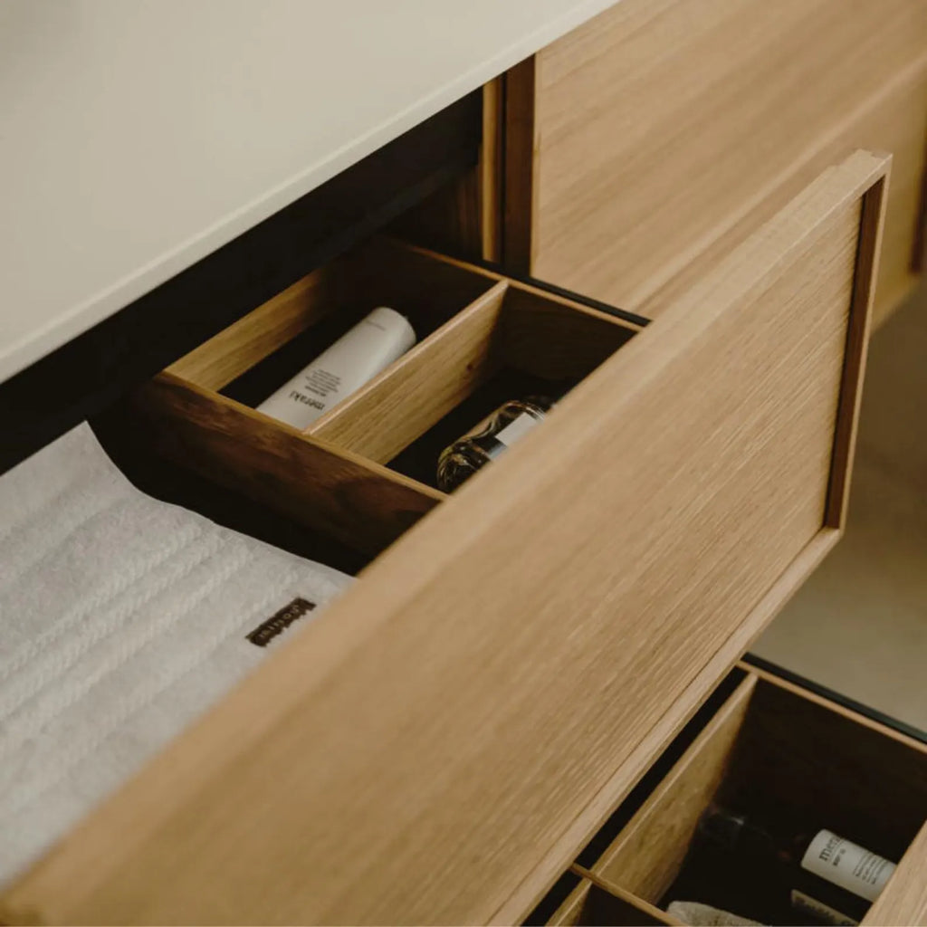 Close-up of a light wood vanity drawer with an open compartment holding toiletry bottles, next to a white towel, in a modern bathroom setting.