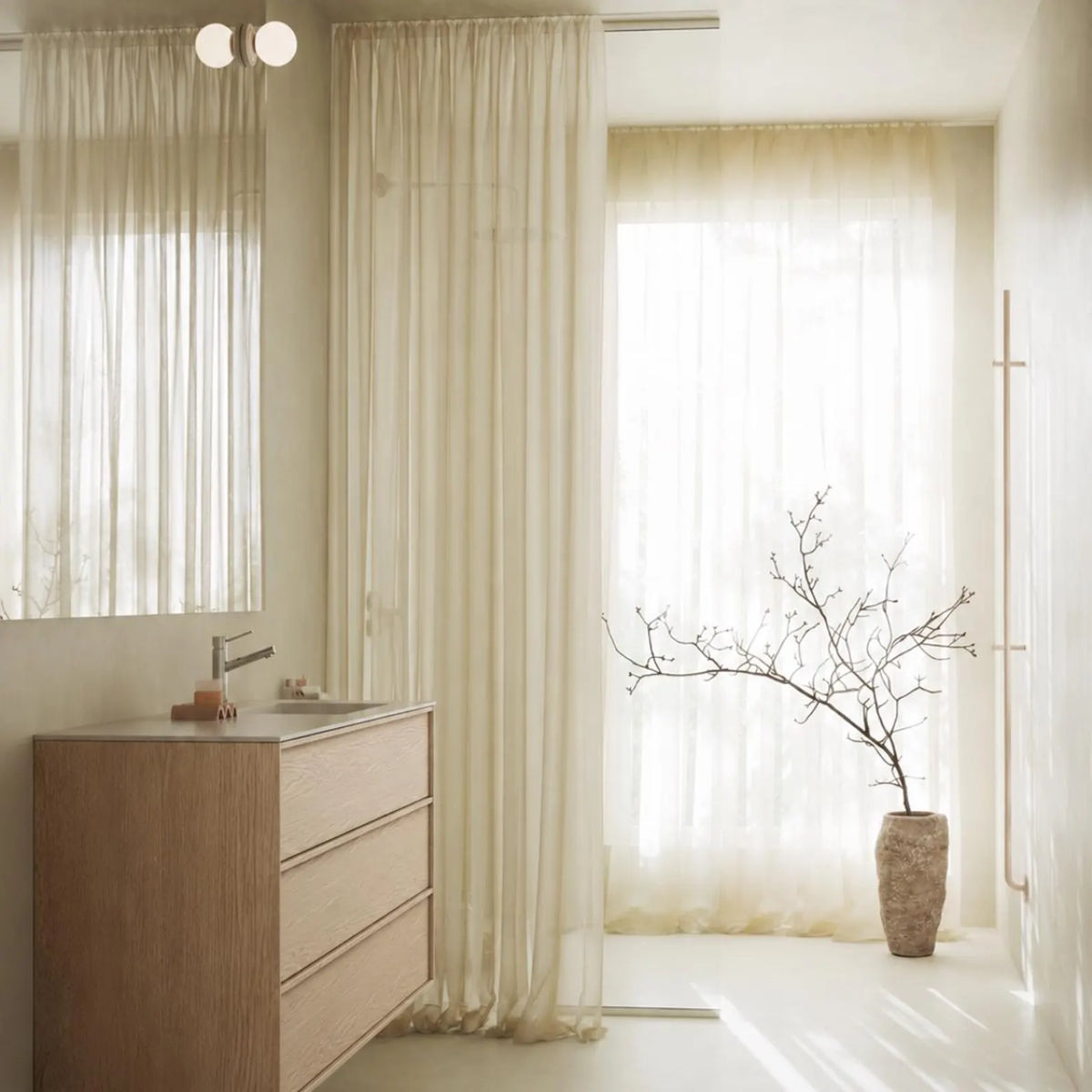 A modern bathroom interior with floor-length sheer cream curtains, a light wood vanity, a large mirror, and a tall textured vase holding bare branches near the window.