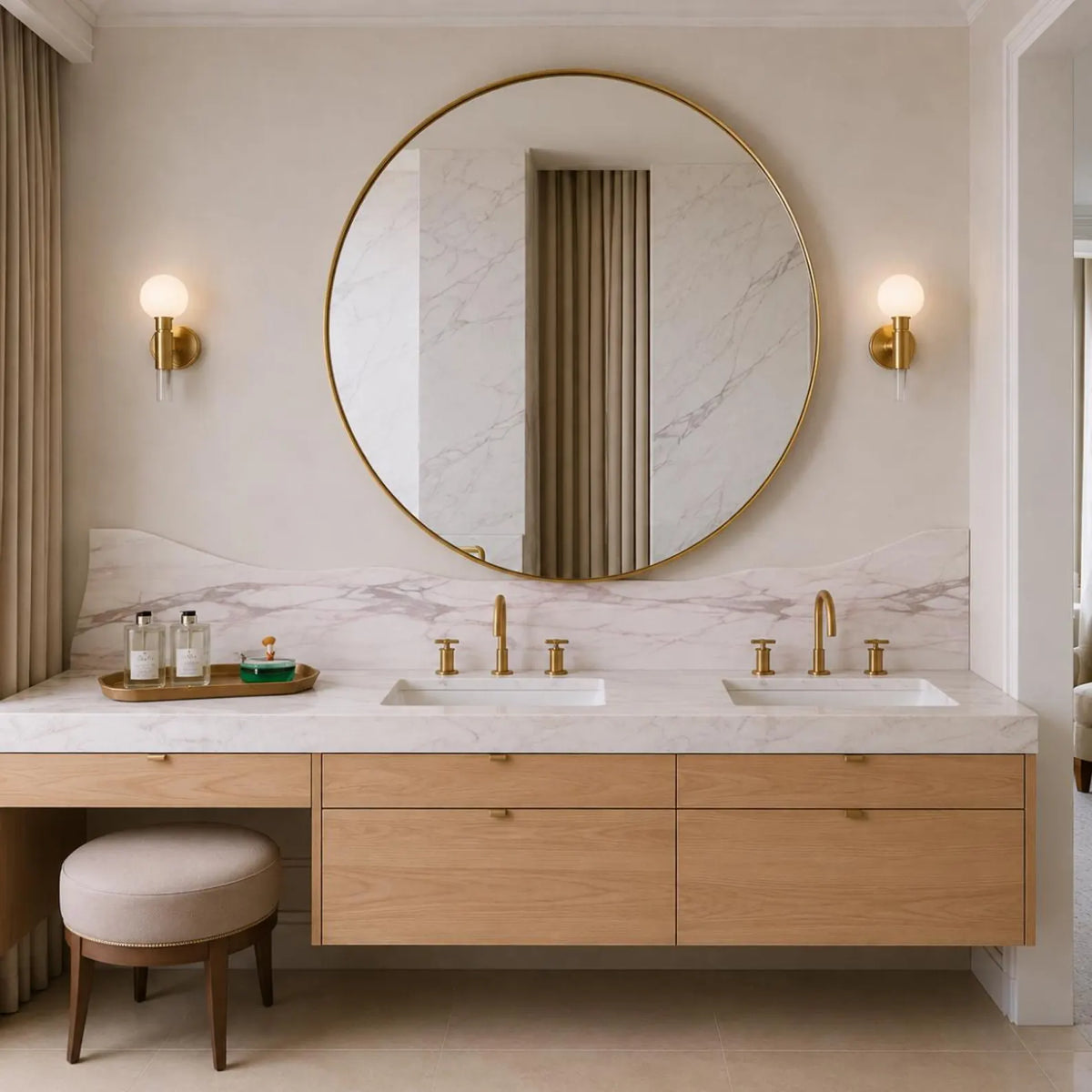 A modern double-sink bathroom vanity with a large round gold-framed mirror, marble backsplash, and light wood floating cabinets. Gold wall sconces, a small stool, and toiletries are visible in a bright, neutral-toned bathroom setting.